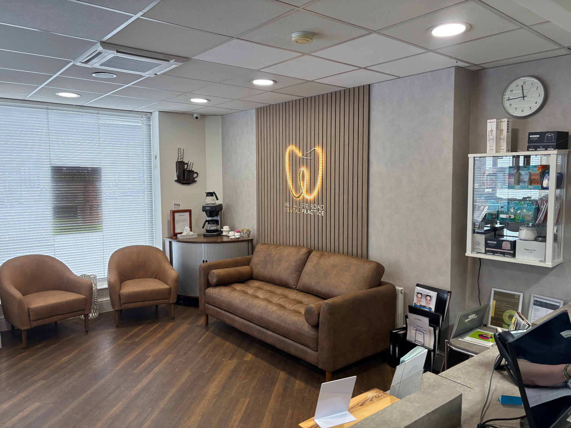 Warm white LED illuminated tooth logo and brushed brass Briercliffe Road Dental Practice lettering on a slatted wood feature wall in the waiting area